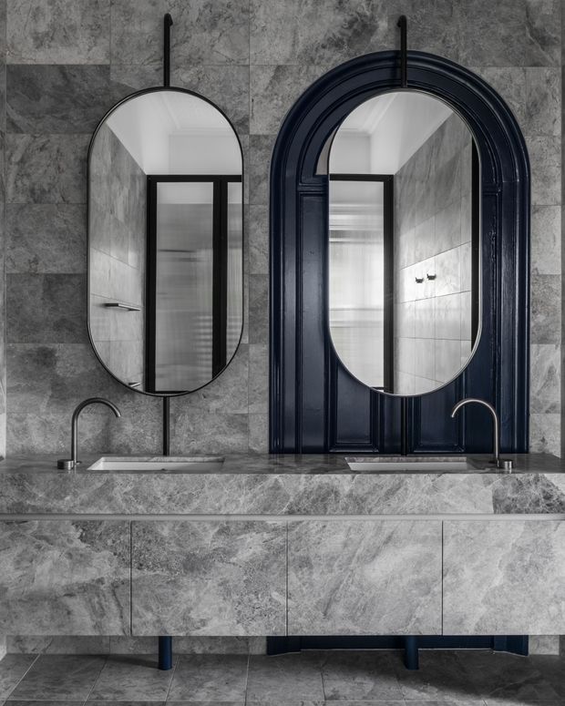 A refined bathroom vanity composition featuring a long grey stone-clad double vanity, two integrated rectangular basins, curved brushed-metal faucets, and paired mirrors. One mirror is an irregular rounded-rectangle form while the other is set within a dramatic dark navy/black arched architectural frame. Grey stone and tile surfaces continue across the walls, benchtop, cabinetry, and floor, creating a monochromatic, high-contrast interior.