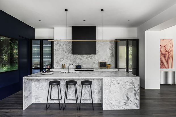 A sophisticated contemporary kitchen dominated by a large marble-clad island with three black bar stools. Matching marble extends across the splashback and rear benchtop, beneath a black rangehood or feature panel. Dark-framed glazing and cabinetry contrast with white walls, dark timber flooring, pendant lights, and black accents. A dining or living area is partially visible beyond the kitchen.