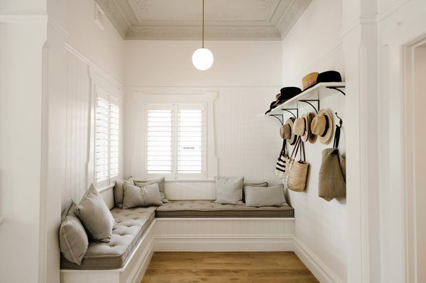 Bright, compact sitting area or sunroom with a built-in L-shaped upholstered bench beneath shuttered windows. The room features white paneled walls, ornate decorative ceiling moulding, a warm timber floor, a simple globe pendant, and wall-mounted open shelving displaying hats and woven bags. The tailored white-and-neutral palette gives it a refined traditional feel.