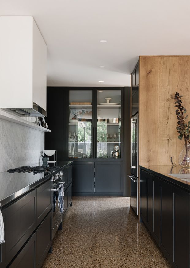 A narrow, galley-style kitchen with dark charcoal cabinetry, a pale stone backsplash, and a warm timber tall cabinet and panelled wall. The kitchen includes a gas cooktop, concealed rangehood, integrated appliances, glass-front display cabinetry, and a polished terrazzo-style floor. Recessed ceiling lights and glazed cabinetry create a refined, contemporary atmosphere.