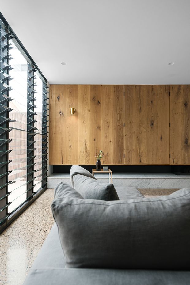 A contemporary living room viewed from behind a large grey sectional sofa. The room features a full-height timber-panelled built-in storage wall, a polished concrete hearth or ledge, expansive black-framed louvred glazing along the left side, recessed ceiling lights, and a speckled terrazzo-like floor. The palette is restrained and warm, combining natural timber, grey upholstery, concrete, and dark metal detailing.