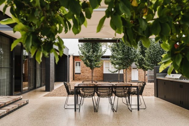A covered outdoor courtyard entertaining area with a large black dining table and wire-frame chairs on a polished light terrazzo or concrete floor. The courtyard is enclosed by warm brick walls and dark framed glazing, with a built-in dark outdoor kitchen or bar along the right side. Mature leafy planting frames the view, and a corrugated metal roof is visible over the adjoining structures.