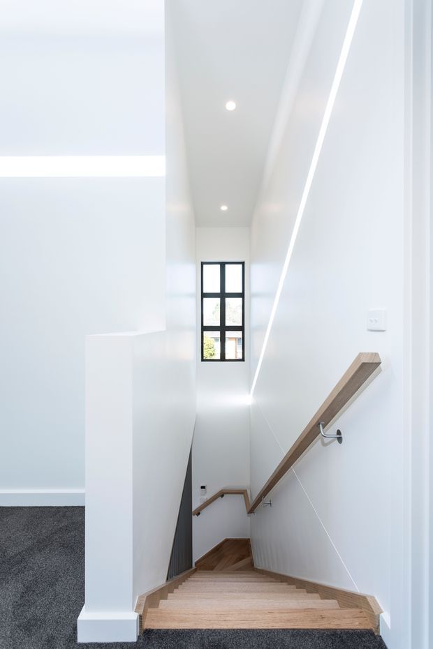 Bright, narrow modern stairwell with white walls and ceiling, a long linear recessed light, dark-framed multi-pane window, and light timber stairs and handrails. The staircase descends between clean white balustrade walls, with dark carpet visible on the adjacent upper landing.