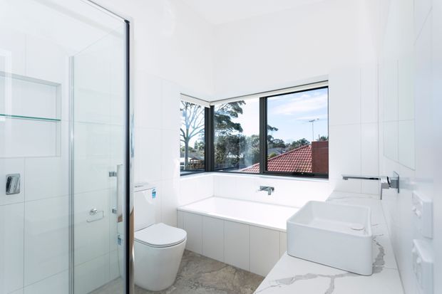 Bright contemporary bathroom with a frameless glass shower enclosure, wall-hung toilet, freestanding bathtub beneath corner windows, and a white vessel basin on a white marble-look vanity. The room has white tiled walls, pale cabinetry, dark window frames, and a light grey stone-look floor. Exterior rooftops and trees are visible through the windows.