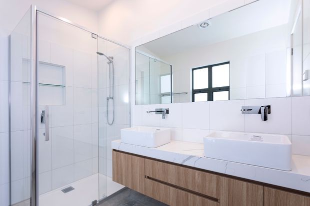Bright contemporary bathroom with a large frameless glass walk-in shower, twin white vessel basins, wall-mounted chrome tapware, a wide timber-look floating vanity, white tiled walls, and a large mirror. A dark-framed window provides natural light, while the darker floor adds contrast.