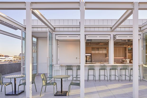 A bright rooftop terrace with a covered outdoor entertaining area, framed by white structural posts and beams. The space includes small tables and chairs, a long bar counter with stools, built-in cabinetry, and a compact outdoor kitchen or serving area. Pale tiled flooring, light architectural finishes, and extensive open glazing create an airy contemporary setting with urban buildings visible beyond.