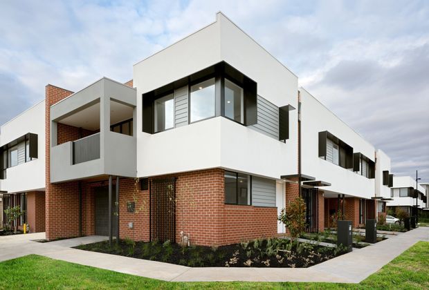 Exterior view of a contemporary multi-unit residential development with crisp white rendered upper volumes, exposed red-brick ground-floor walls, dark-framed corner windows, projecting balconies, and landscaped front paths. The building has a predominantly rectilinear form with flat rooflines and repeated townhouse-like units.