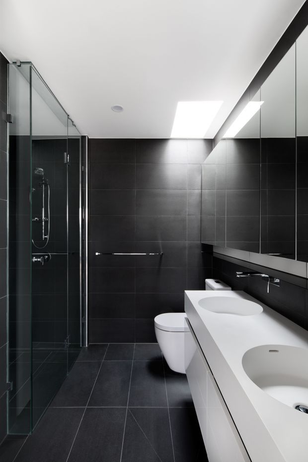 Contemporary bathroom with a long white double vanity and integrated basins, wall-hung toilet, large frameless glass shower enclosure, dark charcoal tiled walls and floor, and a wide mirrored wall. A bright rectangular skylight or ceiling light adds illumination to the otherwise monochrome space.