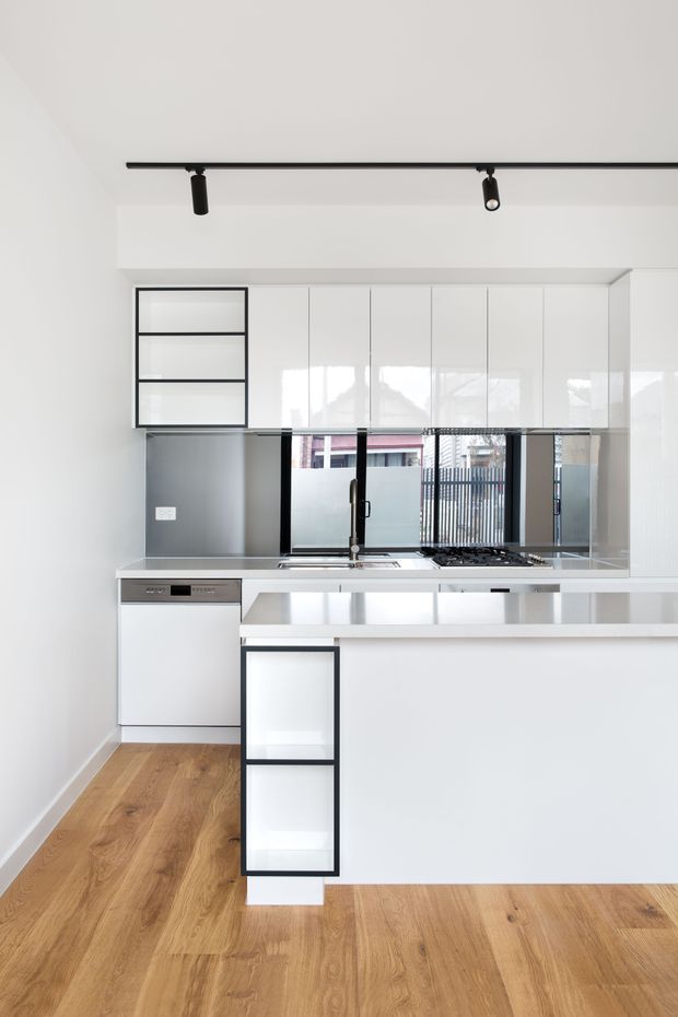 A bright, compact contemporary kitchen with glossy white floor-to-ceiling cabinetry, a white island with integrated open shelving, stainless-steel appliances, a gas cooktop, sink, and reflective splashback. Black-framed open shelving and black track lighting add contrast. Warm timber flooring balances the otherwise white and grey palette, while windows and a glazed door bring in natural light.