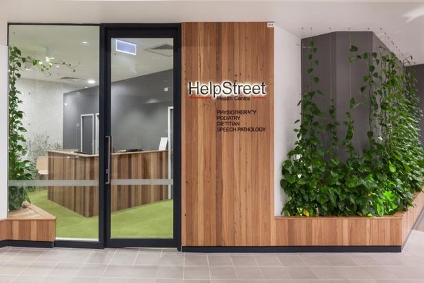 A contemporary healthcare centre reception entrance viewed from a public corridor. The frontage features black-framed glazed doors and sidelights, warm vertical timber wall panelling with HelpStreet signage, integrated timber planter boxes filled with trailing greenery, and a dark grey built-in reception counter visible inside. The interior uses light grey tiled flooring, white ceilings, and recessed lighting.