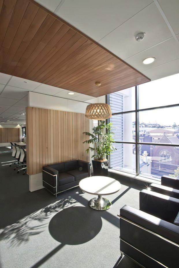 Bright contemporary commercial office lounge and meeting area with black leather armchairs and sofa, a round white coffee table, dark carpet, and a large potted plant. A vertical timber slat feature wall and matching timber-lined ceiling panel add warmth, while full-height black-framed glazing provides abundant daylight and views of surrounding urban buildings. A boardroom or workspace is visible through the opening to the left. A woven pendant light hangs above the seating area.