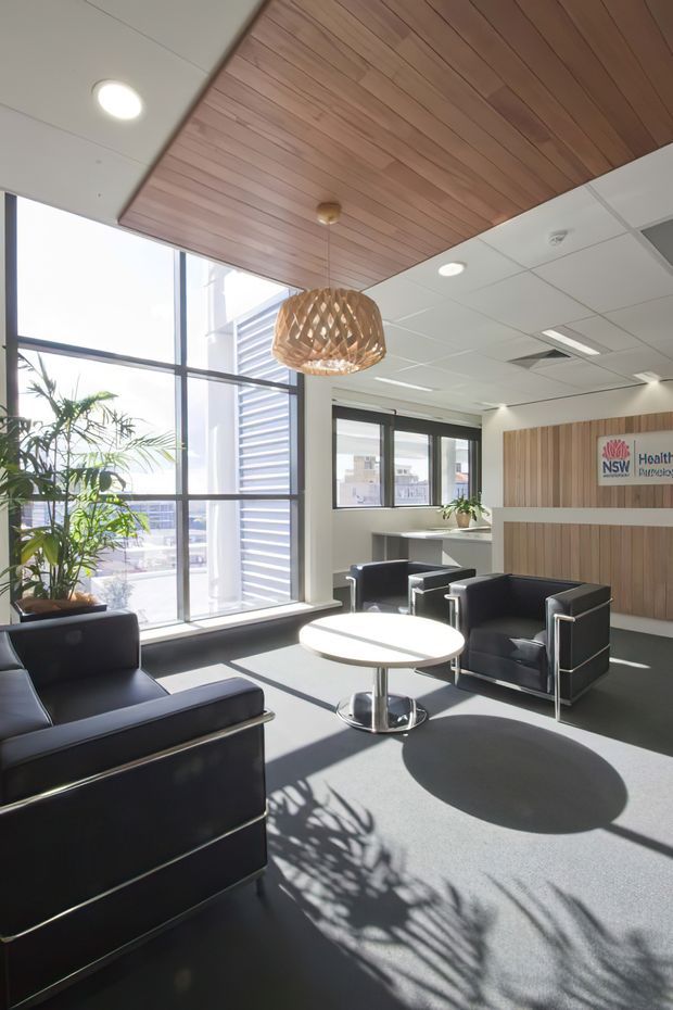 A bright contemporary commercial waiting area or reception lounge with large floor-to-ceiling windows, dark leather armchairs, a round white coffee table, grey carpet, and a timber-lined ceiling feature. A vertical timber wall panel behind the seating includes NSW Health signage, while indoor plants and strong daylight create an open, professional atmosphere.