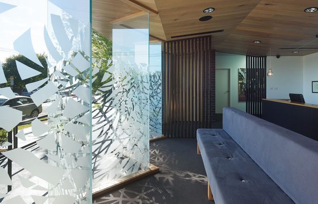 A contemporary reception or waiting area viewed from inside, with a long blue upholstered sofa, dark reception desk, timber-lined ceiling, vertical dark timber screening, and large patterned translucent glass panels. Bright daylight enters from the glazed facade, creating geometric shadows across the carpet and emphasizing the graphic decorative glazing.