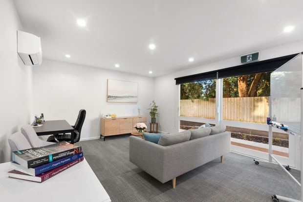 Bright contemporary living room and home office with white walls, recessed ceiling lights, grey sectional sofa, grey area rug, timber media cabinet, compact desk and chair, and large sliding glass doors opening to a fenced outdoor area. A mobile whiteboard stands near the glazing, suggesting a flexible work-from-home setting.
