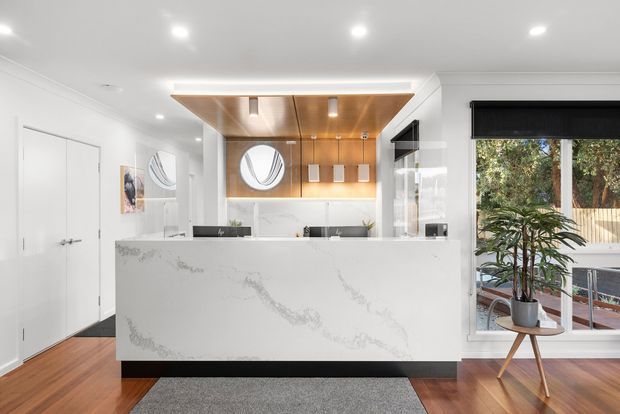 Bright contemporary kitchen with a large white marble-look island, white cabinetry and walls, warm timber flooring, and a striking timber-lined ceiling feature above the cooking and preparation area. A circular window and pendant lights add visual interest, while a large window brings in greenery and natural light.