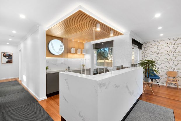 Bright contemporary kitchen with a large white waterfall island, integrated cabinetry, reflective splashback, timber flooring, and a distinctive timber slatted ceiling feature. The open kitchen is adjacent to a hallway and includes a small dining or sitting area with botanical wallpaper.