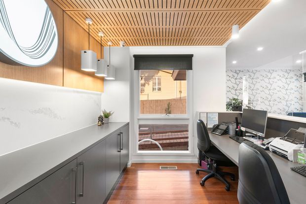 A bright, compact home office with built-in grey cabinetry and a long white benchtop along the left wall, two desk workstations on the right, and a window at the rear. The room features warm timber flooring, a slatted timber ceiling, white walls, pendant lighting, recessed downlights, and a marble-patterned feature wall. A large abstract mirror is mounted above the cabinetry.
