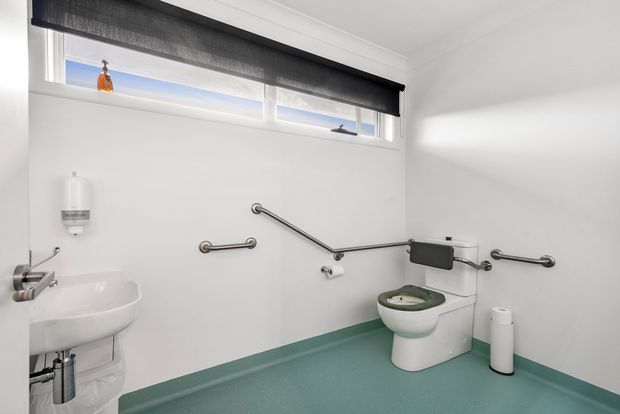 Accessible bathroom with white walls, a mint-green resilient floor, high horizontal windows with a dark roller blind, wall-mounted grab rails, a wall-hung basin, and an accessible toilet with contrasting green seat. The space is bright, functional, and designed for mobility accessibility.