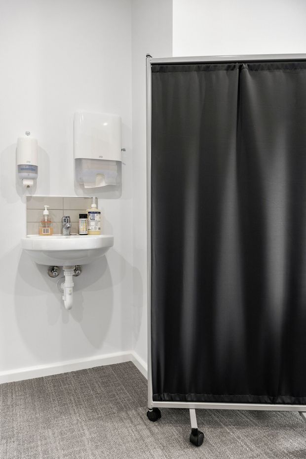 A compact commercial bathroom or clinical wash area with a wall-mounted white hand basin, exposed plumbing, soap dispensers, paper towel dispenser, and a large mobile black privacy screen on casters. The space has white walls, grey carpet flooring, and utilitarian fixtures.