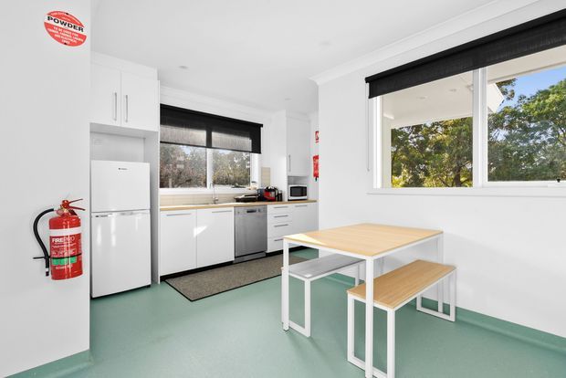 Bright, compact contemporary kitchenette and breakout dining area with white cabinetry, white appliances, a stainless-steel dishwasher, pale timber-and-white table with matching benches, green resilient flooring, and large windows fitted with black roller blinds. A fire extinguisher and powder-room safety signage are visible, suggesting a workplace or commercial staff amenity space.