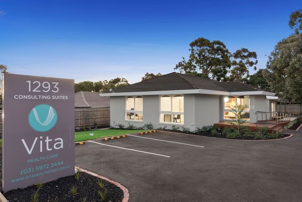 A single-storey healthcare consulting suite with a grey rendered facade, low hipped dark roof, illuminated windows, landscaped edges and a newly paved parking area. A prominent sign identifies Vita Health Care at 1293 Consulting Suites.