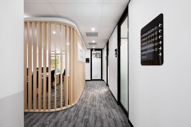 A bright contemporary commercial hallway with grey carpet, white walls, a suspended acoustic-tile ceiling, and black-framed glazed office or consultation-room doors. A curved pale timber vertical-screen partition encloses a waiting or meeting area on the left, while a black directional sign identifies consultation rooms and a toilet.