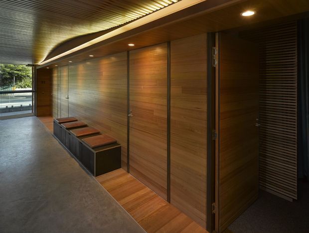A dimly lit contemporary interior corridor with extensive warm timber wall panelling and flush timber doors, dark linear metal detailing, recessed ceiling lights, and a textured metallic ceiling. A long built-in bench or storage unit with dark panels and leather-like seat cushions runs along the wall. The polished concrete floor and open glazing at the end create a refined, minimalist architectural atmosphere.