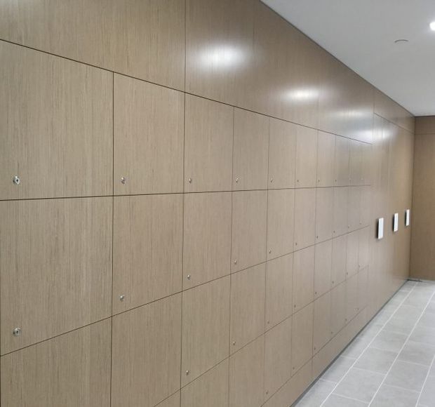 A long, minimalist corridor lined with continuous light timber-look built-in locker cabinetry. The cabinetry has flush rectangular doors with small round metal pulls, extending from wall to ceiling. The floor is finished in large light grey tiles, with a plain white ceiling and recessed lighting.