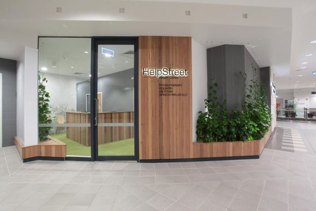 A modern commercial interior in a shopping-centre or medical-office setting, showing a HelpStreet reception and consultation area. The frontage combines black-framed glazed doors and partitions with warm vertical timber wall cladding, grey built-in walls, integrated planter boxes with greenery, and a pale tiled floor. Recessed ceiling lighting and an open corridor extend into the background.