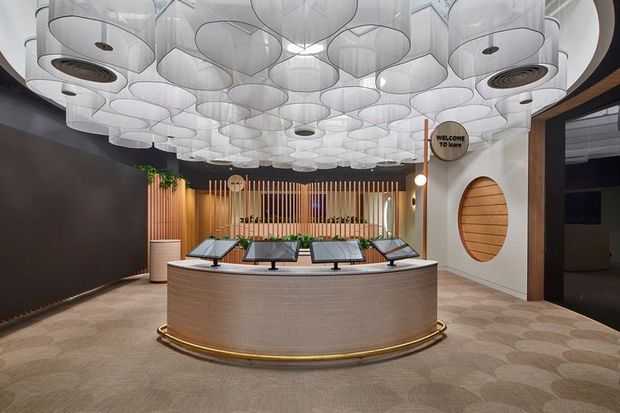 A refined commercial reception or concierge lobby featuring a large curved, textured reception desk with brass trim, multiple angled display screens, a dramatic canopy of translucent sculptural pendant lights, timber screening with integrated greenery, and a rounded feature wall with a timber-lined oval inset. The palette is warm and sophisticated, with dark walls, timber details, cream surfaces, and patterned carpet.