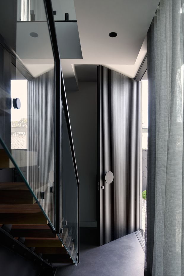 A contemporary interior view of a compact stairwell and entry transition. Dark timber stair treads rise beside a clear glass balustrade with black metal framing, while a tall black vertically ribbed feature wall and matching pivot door dominate the space. The ceiling is white with recessed downlights and concealed linear lighting, and sheer grey curtains filter daylight at the right. The image also captures reflections in the glazing and a glimpse of landscaped exterior beyond the open door.