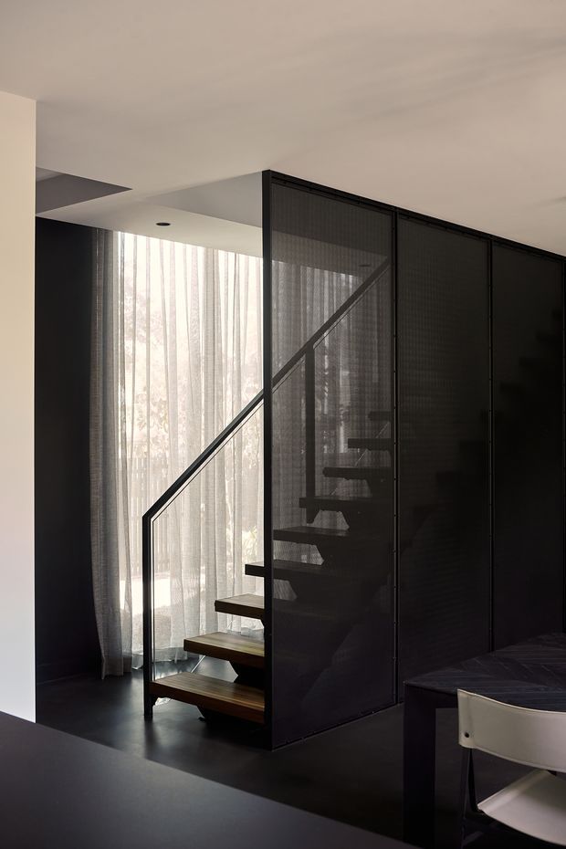 A dark, contemporary interior featuring an open-riser timber staircase enclosed by a black metal-framed mesh screen. Sheer floor-to-ceiling curtains filter daylight beside the stair, while a dark dining table and light upholstered chair are partially visible in the foreground. The space has a restrained, graphic palette and minimal detailing.