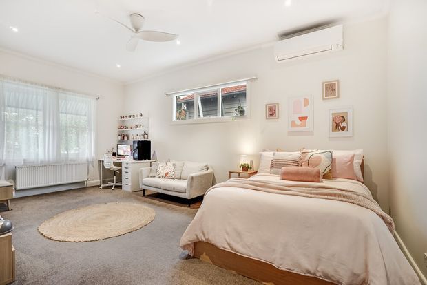 A bright, softly styled bedroom with a large upholstered bed, layered blush and cream bedding, a compact seating area, grey carpet, sheer-curtained window, white walls, ceiling fan, wall-mounted air conditioner, and delicate framed artwork. The room has a calm, warm, contemporary residential interior.