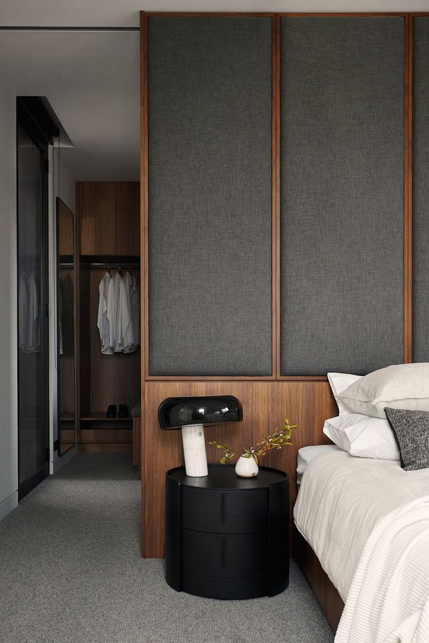 A refined bedroom interior with a partially visible upholstered bed, white bedding, and a dark textured accent cushion. The room features a full-height timber and charcoal fabric wall panel behind the bed, integrated wardrobe storage with hanging clothes, dark carpet, and a compact black bedside table with a contemporary lamp and small vase. The palette is restrained and warm, combining walnut-toned timber, charcoal grey, black, and off-white.