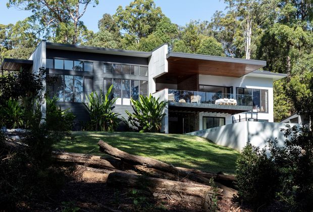 Exterior view of a contemporary multi-level home set among mature trees on a sloping, landscaped site. The house features crisp white rendered forms, extensive dark-framed glazing, projecting flat roof planes, a timber-lined soffit, and a glass-balustraded upper terrace or balcony.