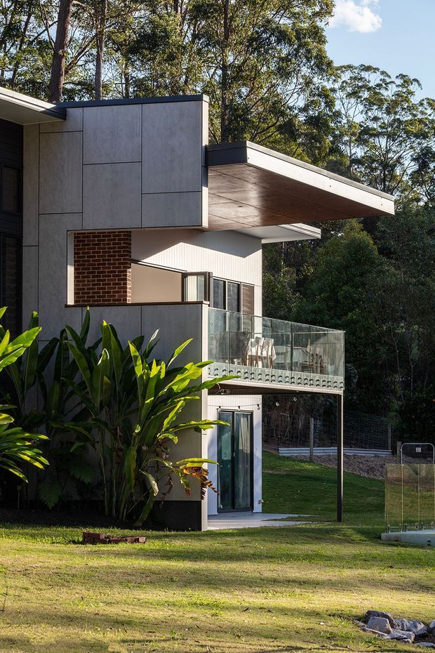 Exterior view of a contemporary multi-storey home set among mature trees and landscaped grounds. The house features flat projecting roof planes, smooth grey concrete wall panels, exposed red brick, large glazed openings, and a glass-balustraded balcony with a covered outdoor area below.