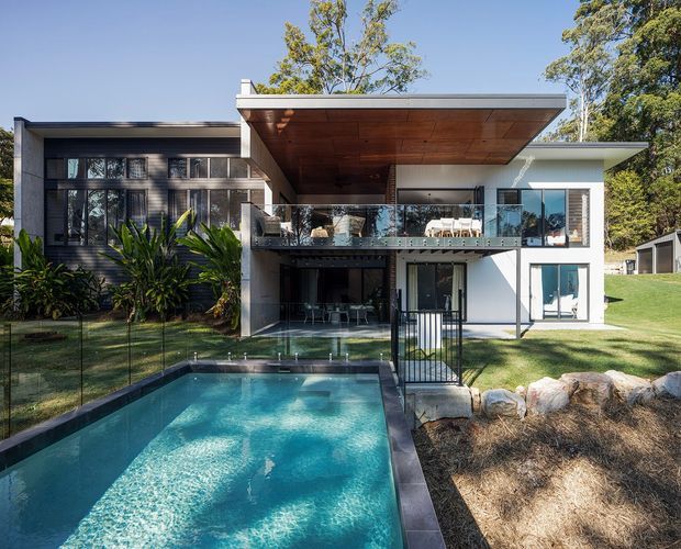 Exterior view of a contemporary multi-level home with crisp white and dark charcoal facade elements, extensive glazing, a broad cantilevered upper-level roof and warm timber soffit. A balcony and covered outdoor living area overlook a rectangular swimming pool, with landscaped planting and mature trees surrounding the property.