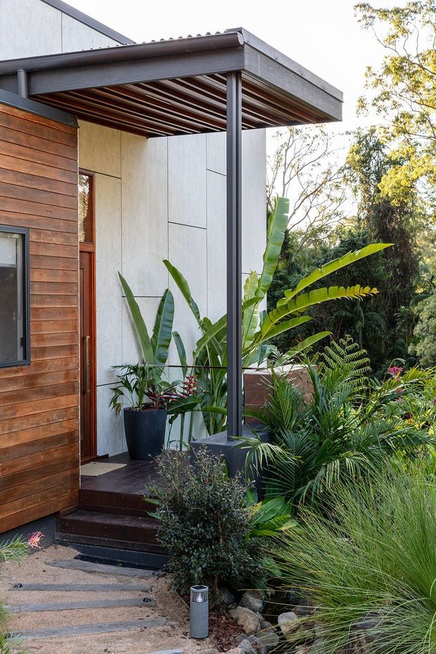 Exterior view of a compact contemporary home entry and covered porch, featuring horizontal timber wall cladding, pale fibre-cement or concrete panel walls, dark metal posts and beams, a slatted timber soffit, and dark timber decking steps. Lush tropical and ornamental planting surrounds the walkway and porch.