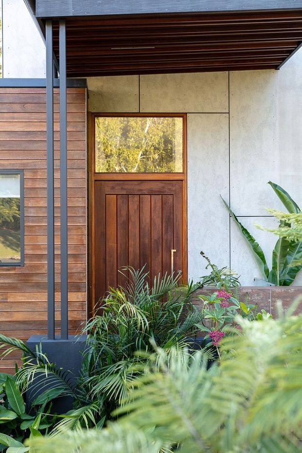 Close-up exterior view of a residential entry featuring a richly stained timber door with a glazed upper panel, horizontal timber wall cladding, pale concrete or fibre-cement wall panels, dark slender metal supports, and a timber-lined covered porch ceiling. Dense tropical planting softens the foreground.