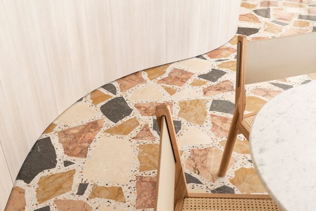 Close-up interior view of a curved light-wood built-in or wall panel beside a terrazzo-style floor with large irregular stone chips in cream, terracotta, tan, and dark grey. A wooden dining chair and the edge of a round pale stone or terrazzo tabletop are visible, with a matching irregular-stone wall surface in the background. The image has a warm, refined contemporary feel.