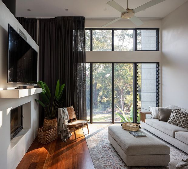 A bright contemporary living room with a double-height glazed wall overlooking greenery. The room features a large sectional sofa and ottoman, a wall-mounted television above a sculptural fireplace, dark full-height curtains, timber flooring, a woven planter, and a large ceiling fan. Natural light creates a warm, inviting atmosphere.