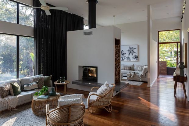 A bright, open-plan living room with rich dark timber flooring, a central white fireplace volume, full-height glazing, and black curtains. The foreground includes a sofa, woven rattan lounge chairs, and round timber coffee tables, while a secondary sitting area is visible beyond the fireplace. Tall windows overlook leafy surroundings, and a glazed door opens to an exterior balcony or terrace. A ceiling fan, pendant lights, built-in firewood storage, and framed artwork complete the relaxed contemporary interior.