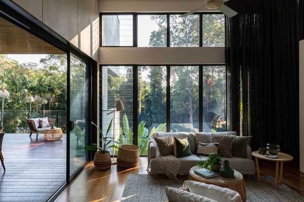 A bright, double-height living room with extensive black-framed floor-to-ceiling glazing overlooking lush vegetation. The room features warm timber flooring and wall surfaces, a neutral upholstered sofa with layered cushions and throws, a woven coffee table, indoor plants, and a large ceiling fan. A glazed connection opens to a timber outdoor deck with additional seating. Dark vertical drapery creates contrast along one side of the room.