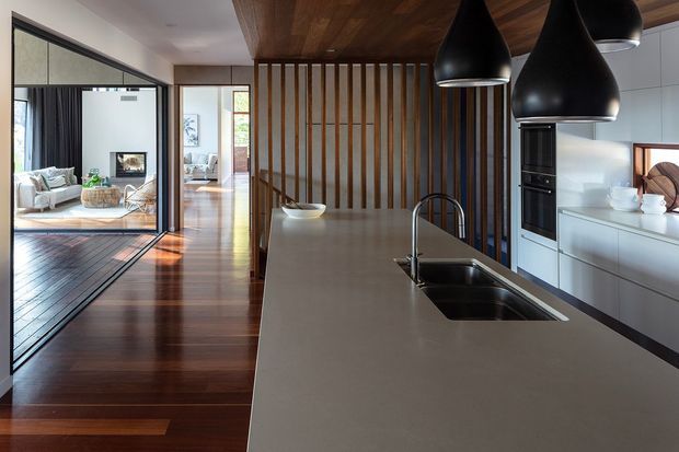 Contemporary open-plan kitchen with a large pale island bench, integrated double sink, white cabinetry and black built-in appliances. Three oversized black pendant lights hang beneath a warm timber ceiling, while a vertical timber slat screen separates the kitchen from the timber-floored living area. Black-framed full-height glazing and sliding doors provide a visual connection to the adjacent living room and exterior.