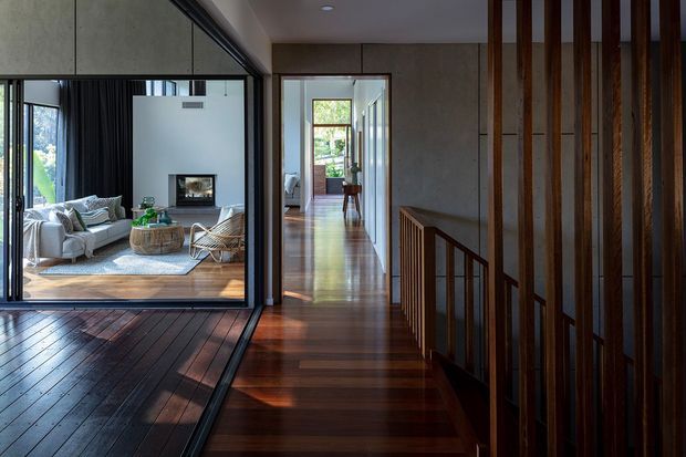 Interior view of a contemporary timber-lined hallway and landing, with dark polished timber flooring, a timber stair balustrade, and vertical timber screening. A glazed opening on the left looks into a bright living room with a sofa, fireplace and woven coffee table. The corridor extends toward a sunlit room and garden-facing glazing.