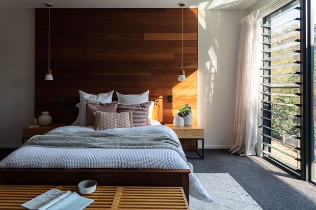 A contemporary bedroom with a low platform bed positioned against a dark timber feature wall. Layered neutral and patterned bedding, pendant lights, bedside joinery, and a slatted timber bench create a warm, refined interior. Full-height louvred glazing on the right brings in strong natural light and views of foliage.