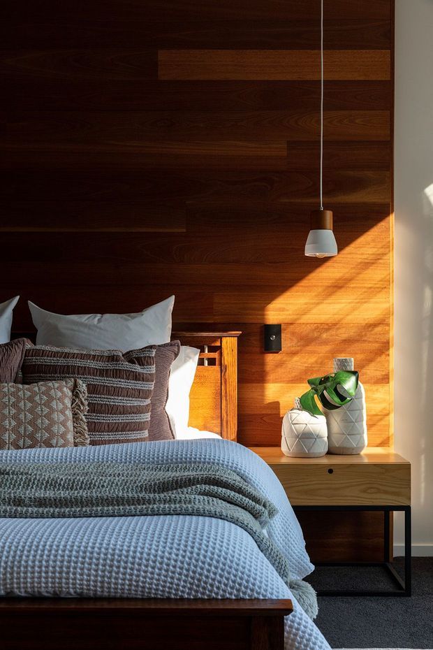 A warmly lit bedroom featuring a bed with layered blue-grey textured bedding and patterned cushions, set against a dark timber feature wall. A timber bedside table with black metal legs, pendant light, and decorative white vases with greenery complete the composed interior.
