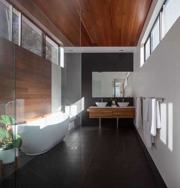 A contemporary bathroom with a freestanding white bathtub enclosed by clear glass, a dark tiled feature wall and floor, twin white vessel basins on a floating timber vanity, and a large horizontal mirror. Warm timber ceiling and wall panelling contrast with the dark finishes, while high windows provide strong natural light. A towel rail and indoor plants complete the refined, spa-like space.