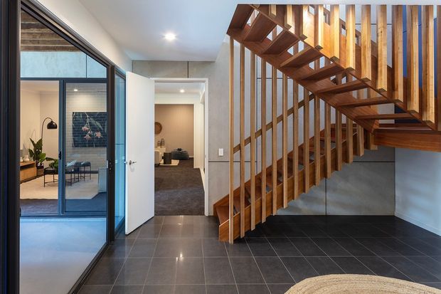 Interior view of a contemporary entry hall dominated by an open-riser timber staircase with vertical timber balusters and a matching timber balustrade. Large black-framed glazed sliding doors reveal an adjacent styled room, while a tiled floor, white walls, and recessed ceiling lighting create a clean, refined scheme.