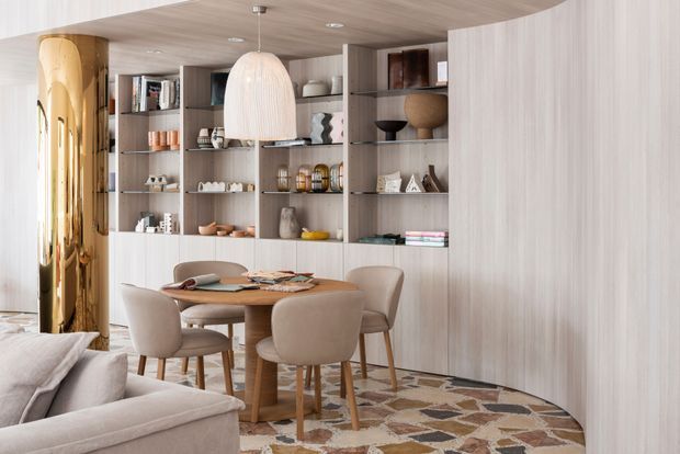 A refined open-plan dining area adjoining a living room, with a round timber dining table and upholstered chairs set in front of extensive built-in open shelving. The shelving displays ceramics and decorative objects, while a large ribbed pendant light hangs over the table. Curved timber-look ceiling and wall panelling, a glossy bronze-toned curved feature doorway, and a patterned stone or tile floor create a warm, contemporary interior.